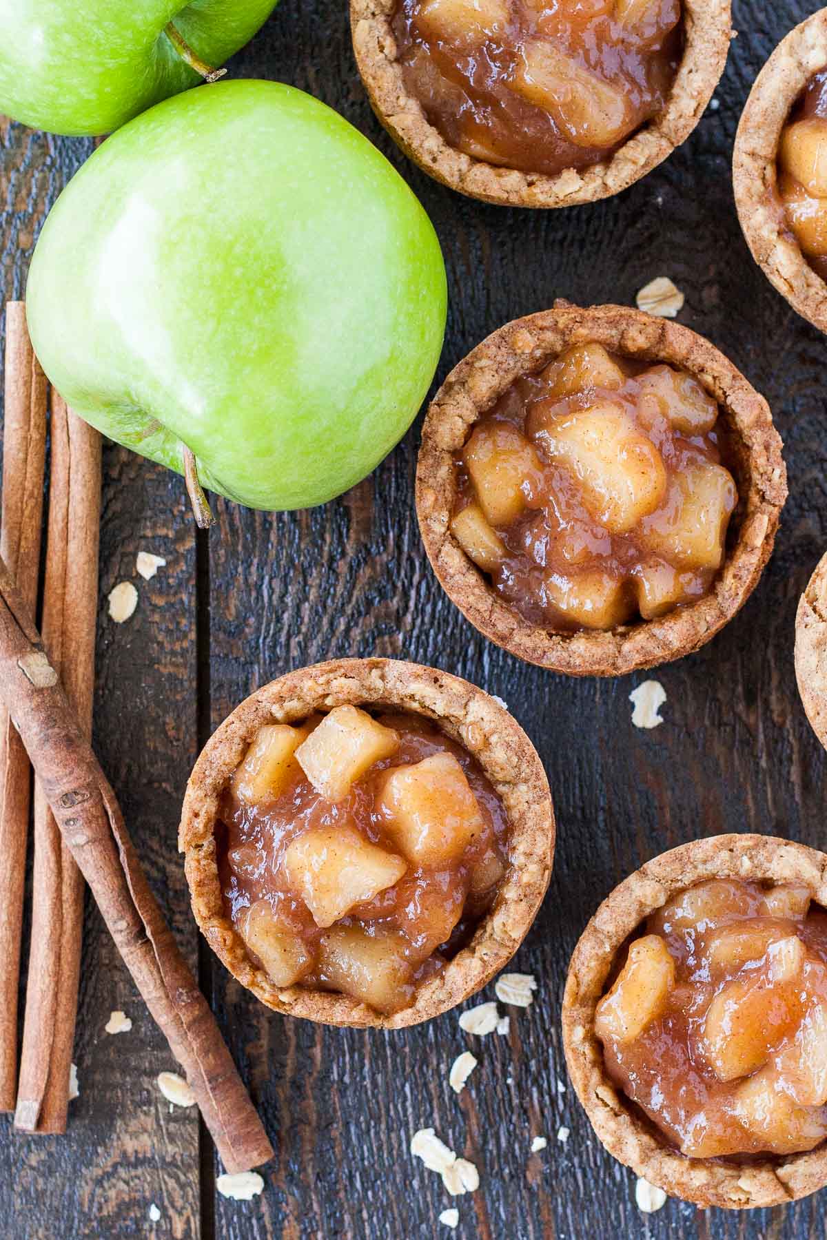 These Apple Crisp Cookie Cups combine classic oatmeal cookies with homemade apple pie filling for the perfect comfort food. | livforcake.com Overhead photo of apple crisp cookie cups with apples and cinnamon sticks on the side.