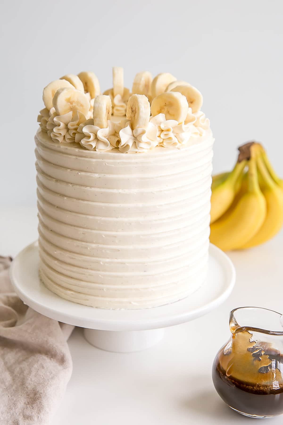 Bananas Foster cake with bananas in the background and fosters sauce in a small pitcher.