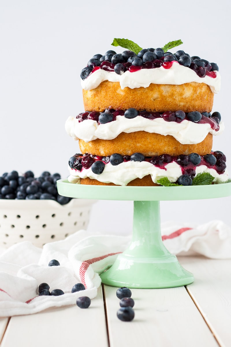 This Blueberry Shortcake Cake is the perfect light dessert for summer. Layers of vanilla cake, whipped cream, blueberry sauce, and fresh blueberries. | livforcake.com Blueberry shortcake decorated in a rustic, naked cake style on a green cake stand with a bowl of blueberries to the side.