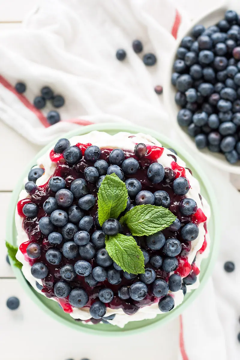 This Blueberry Shortcake Cake is the perfect light dessert for summer. Layers of vanilla cake, whipped cream, blueberry sauce, and fresh blueberries. | livforcake.com Overhead shot of the cake with the fresh blueberries piled on top. A white tea towel to the side.
