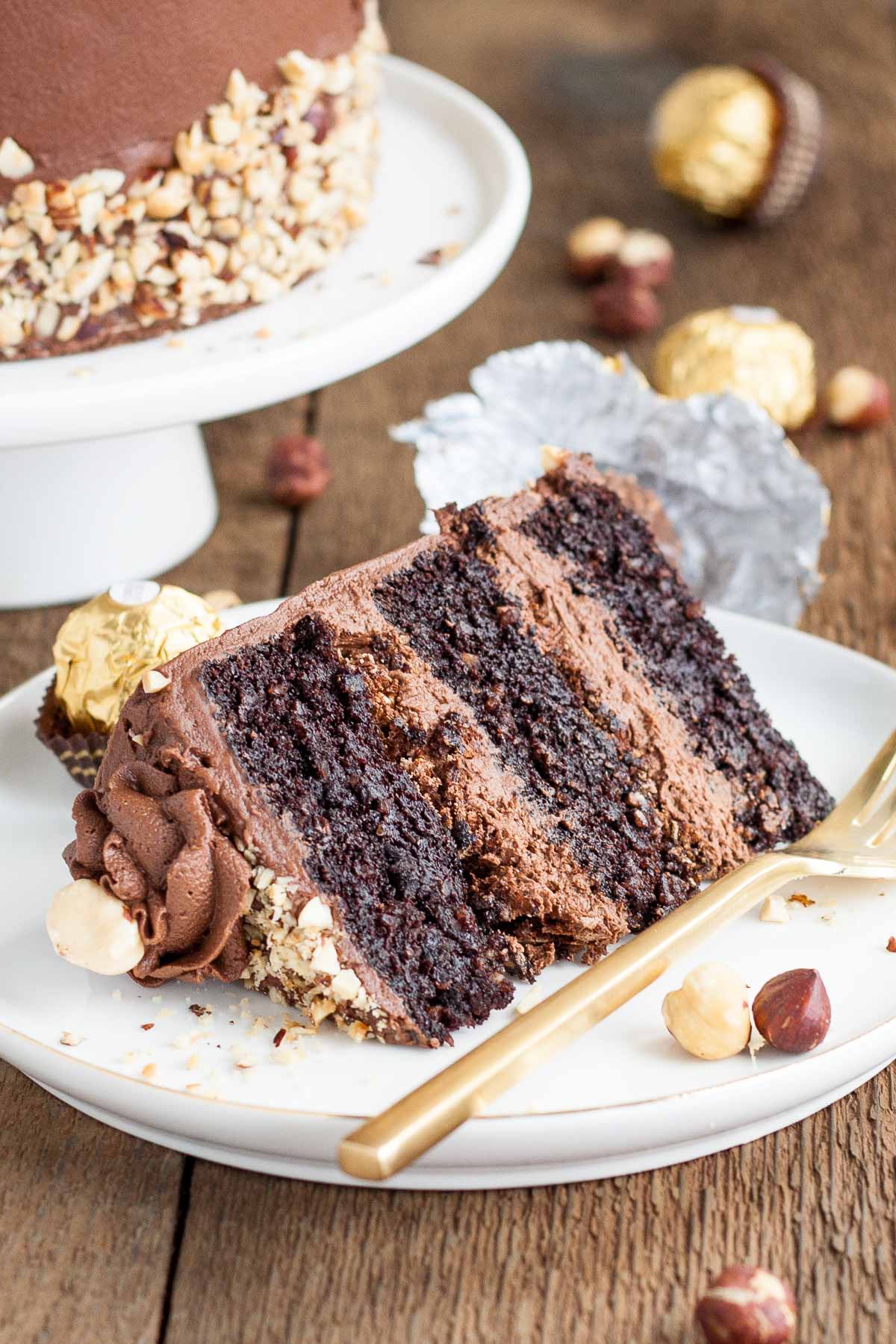 This Ferrero Rocher Cake is your favourite chocolate hazelnut treat in cake form! Chocolate hazelnut cake layers and with a Nutella buttercream. | livforcake.com Slice of Ferrero Rocher Cake on a plate with a gold fork.
