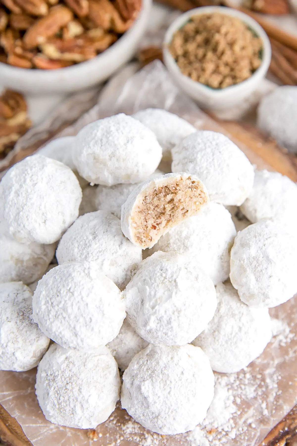 These Mexican Wedding Cookies (also known as Snowball Cookies or Russian Tea Cakes) are buttery, melt-in-your-mouth delicious! Pecan cookies with a hint of cinnamon dusted with powdered sugar. | livforcake.com Stacked cookies with pecans in the background.