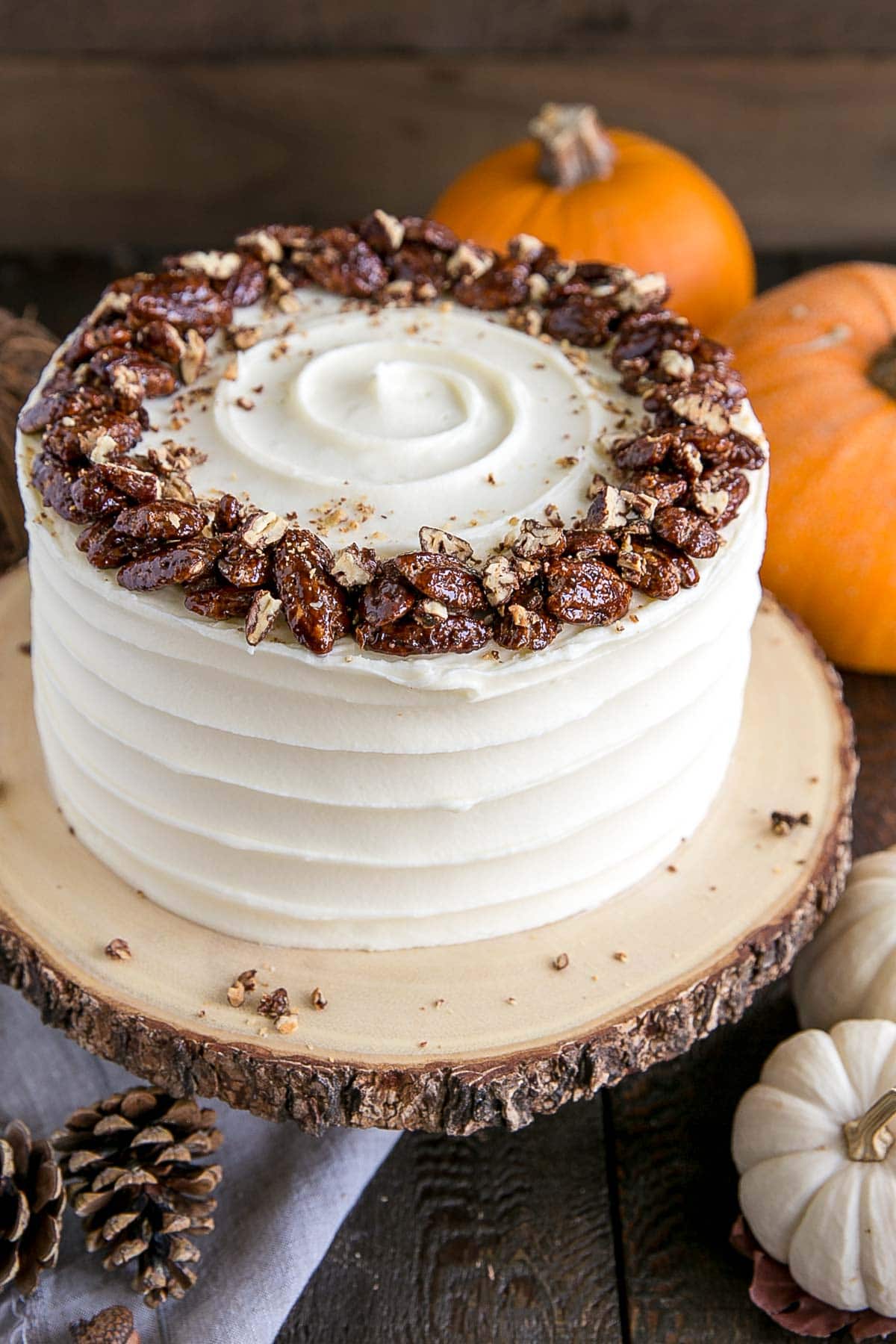 This pumpkin cake is perfect for Fall! Moist and delicious pumpkin cake layers with a tangy cream cheese frosting and crunchy candied pecans. | livforcake.com Cake on a rustic cake stand with pumpkins in the background.